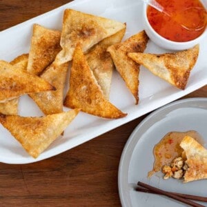 several completed tofu wontons on a white serving platter with a sauce on the side against a brown background