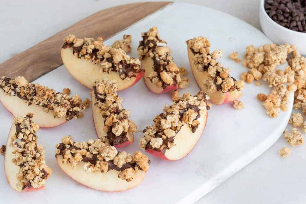 several apples with granola and chocolate on top on a white cutting board