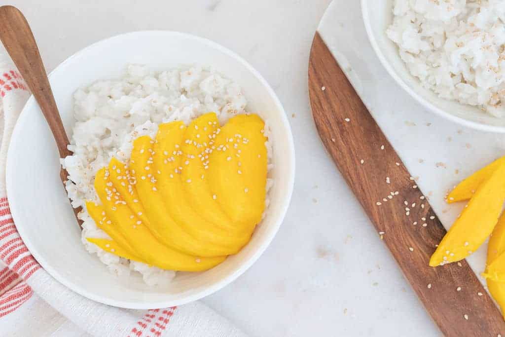 completed mango sticky rice in a white bowl with a bowl of rice and mangos in the background