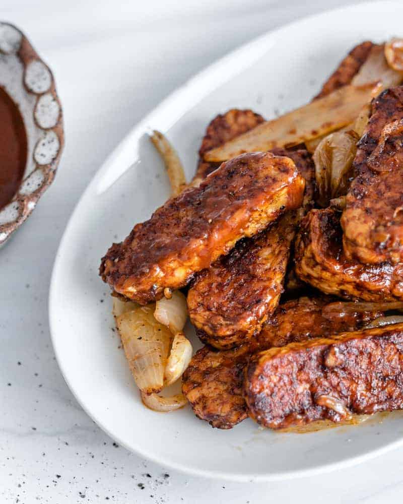 completed bbq tempeh plated on a white plate with BBQ sauce on the side