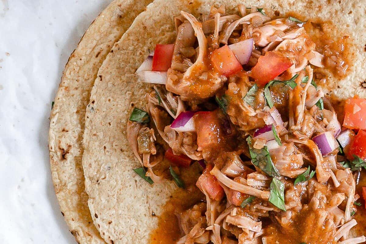 completed jackfruit carnitas on tortillas against a white background with ingredients in the background
