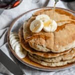 completed stack of banana pancakes on a white plate with bananas and maple syrup on top