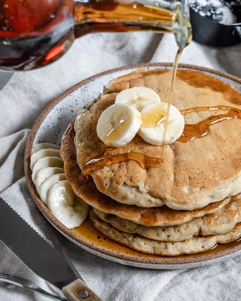 completed stack of banana pancakes on a white plate with bananas and maple syrup on top