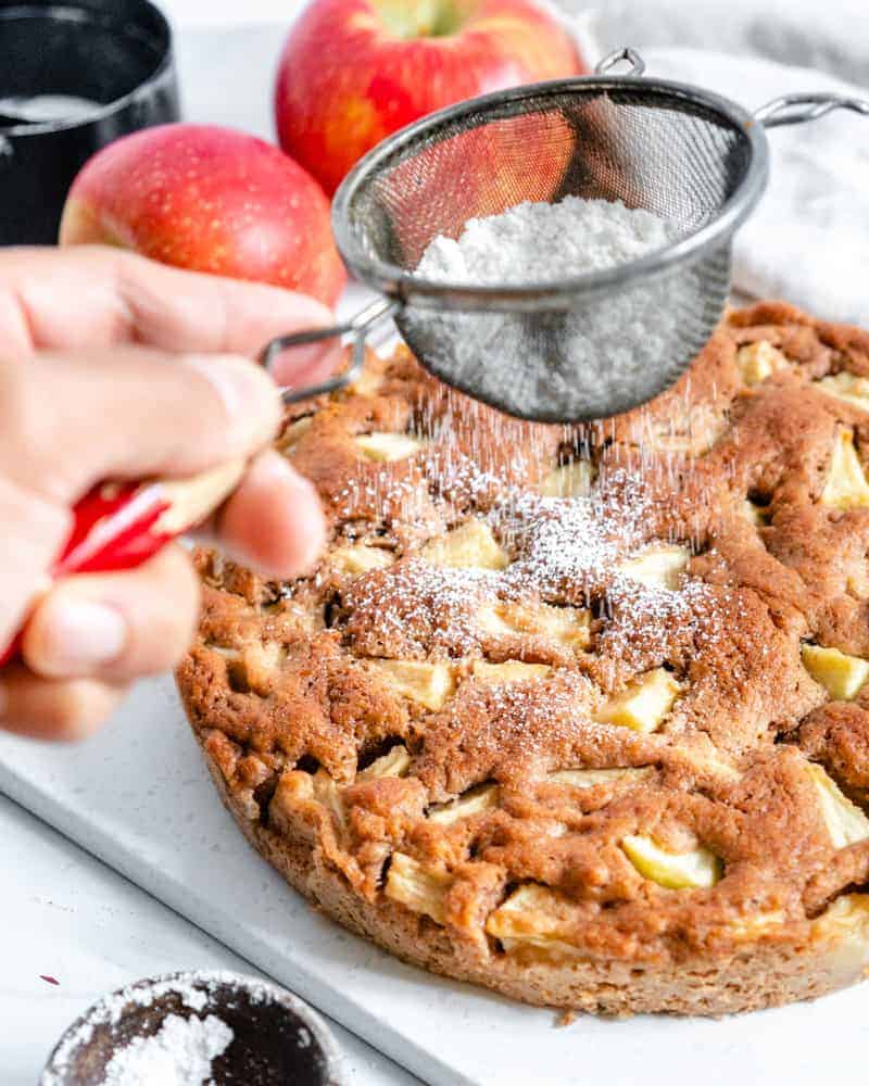 finished apple cake with two apples and measuring tools against a white background and sugar being sprinkled on top