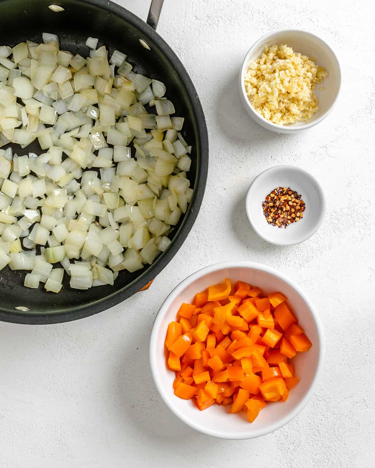 onions in a black pan alongside small bowls of veggies against a white surface