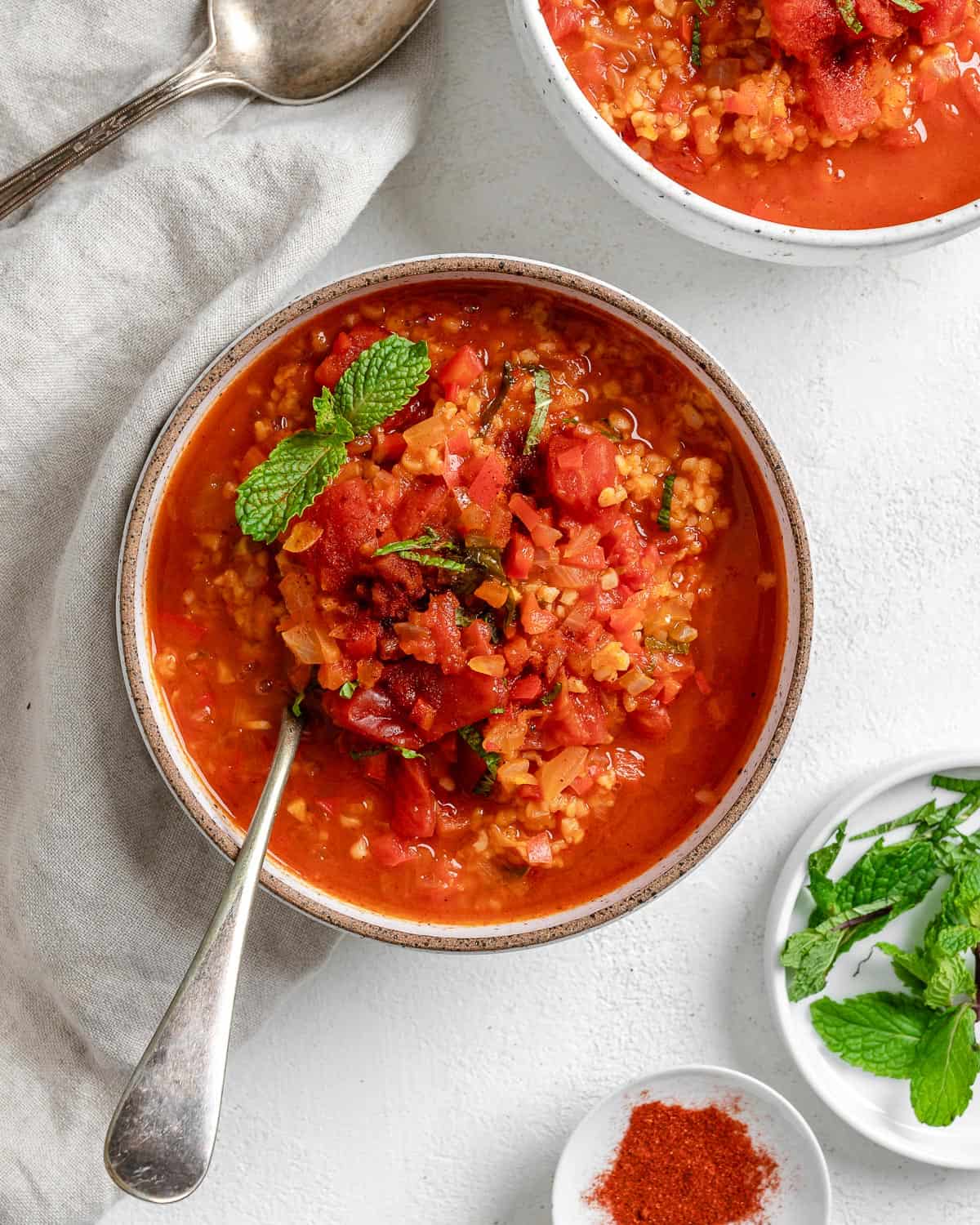 completed Turkish Tomato, Bulgur, and Red Pepper Soup in a bowl against a white surface with herbs and extra soup in the background
