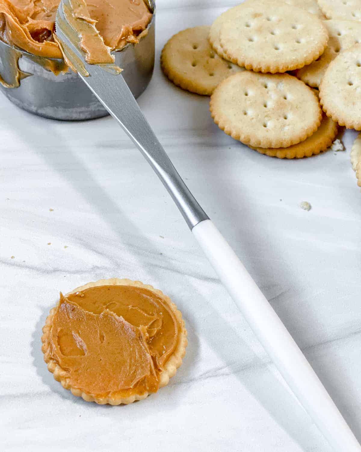 process of making Peanut Butter Cracker Sandwiches and spreading peanut butter on crackers against white surface