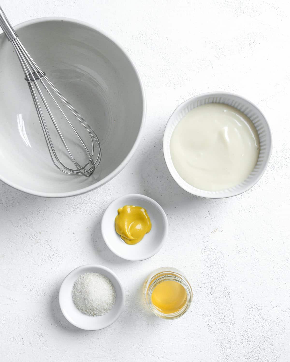 measured out condiments in small white bowls alongside mixing bowl and mixer utensil against white surface