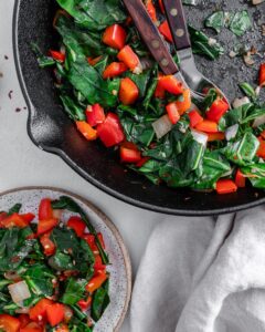 completed Spicy Collard Greens in a black pan against a white surface with a serving in a clear container on the side