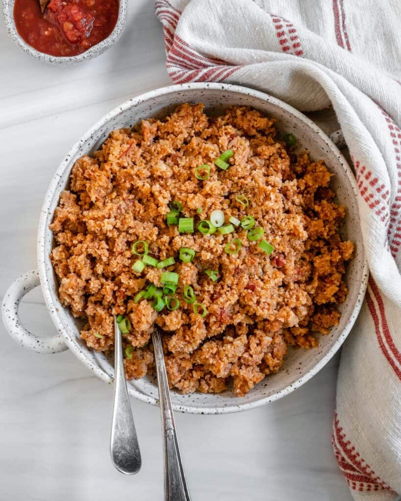 completed Spanish Cauliflower Rice in a white bowl against a white surface with two utensils in the bowl