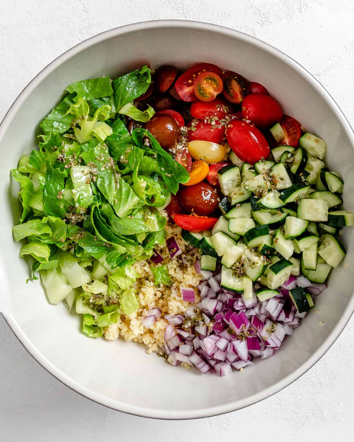 process of adding ingredients for Vegan Greek Millet Salad in a white bowl against a white background