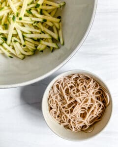 sliced cucumber in bowl and soba noodles in separate bowl against white surface