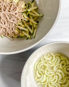 sliced cucumber mixed with soba noodles in white bowl with another bowl of cucumbers in marinade