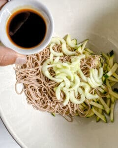 process of adding soy sauce to cucumber and soba noodles bowl