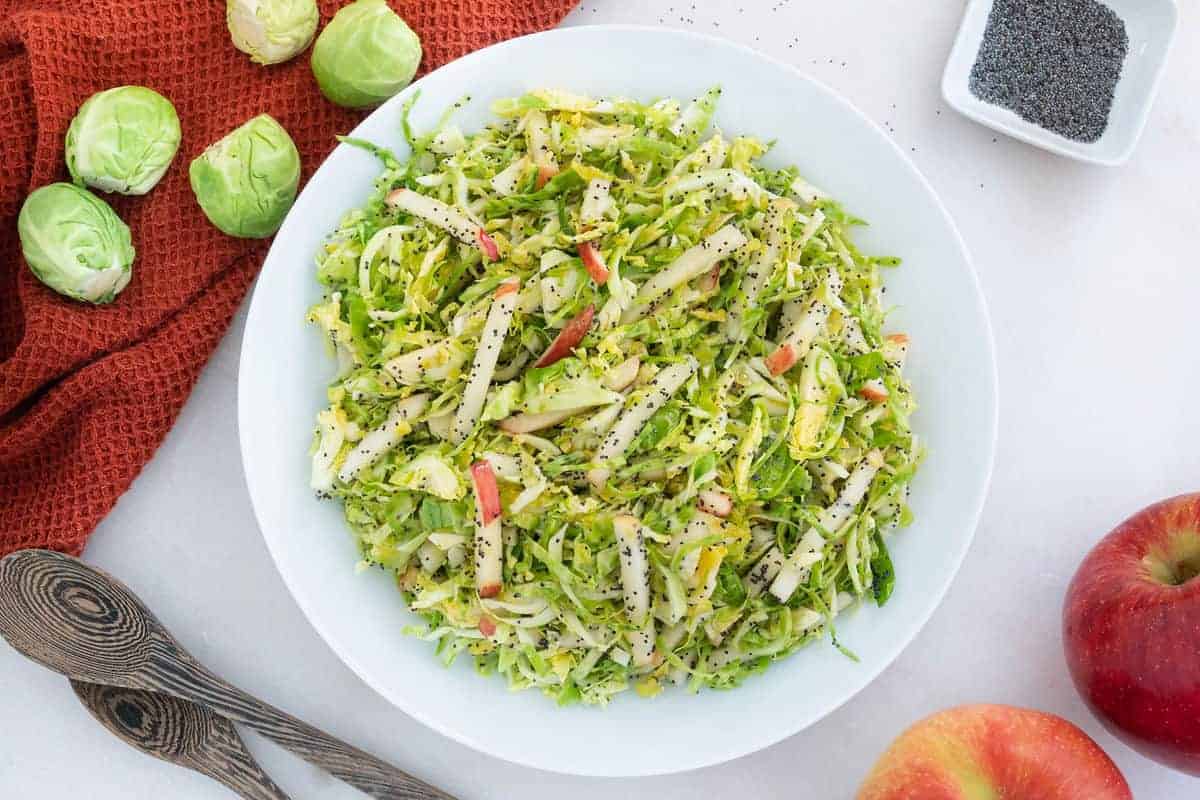 Brussels Sprouts Slaw in a white bowl  with apples and brussels sprouts on the side