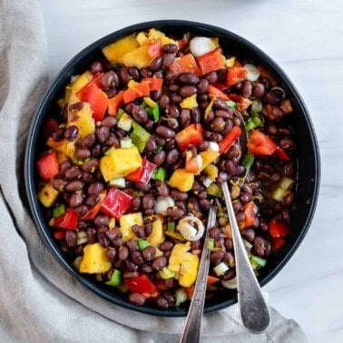 completed mango bean salad in a dark bowl with utensils in the bowl against a white background