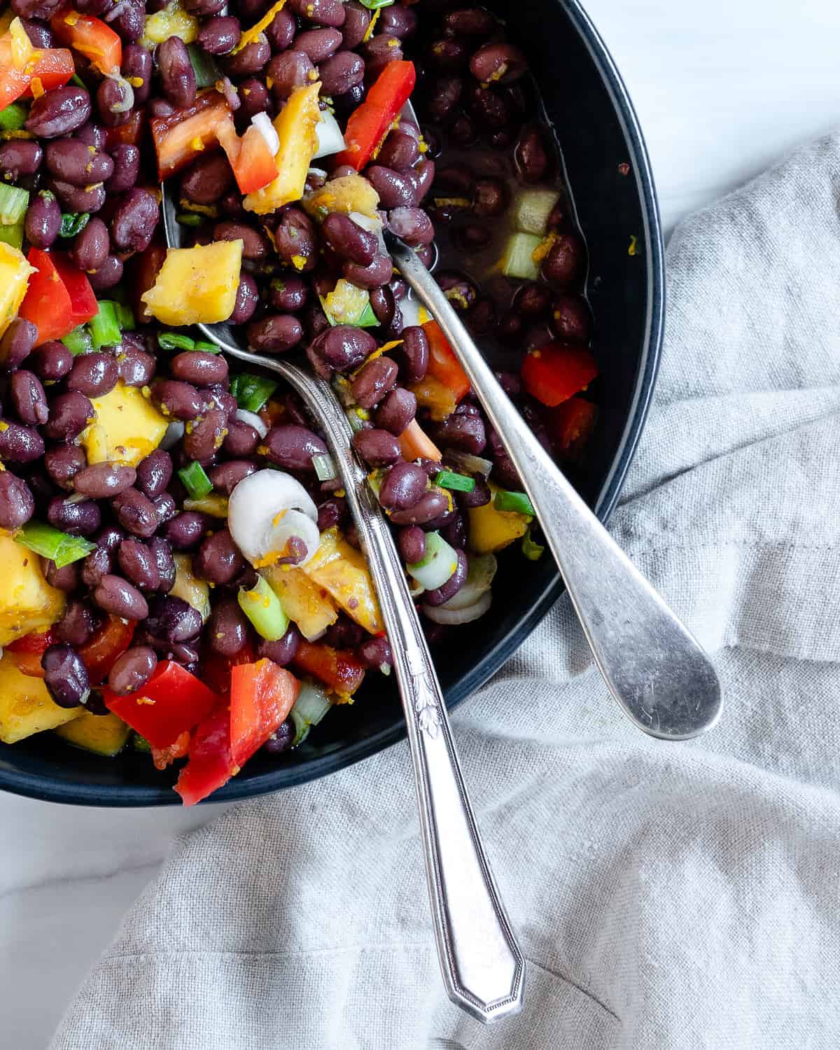 completed mango bean salad in a dark bowl with utensils in the bowl against a white background