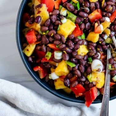 completed mango bean salad in a dark bowl with utensils in the bowl against a white background