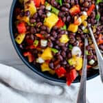 completed mango bean salad in a dark bowl with utensils in the bowl against a white background