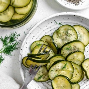 completed Marinated Cucumber Salad on a white plate with cucumber in the background against a white surface