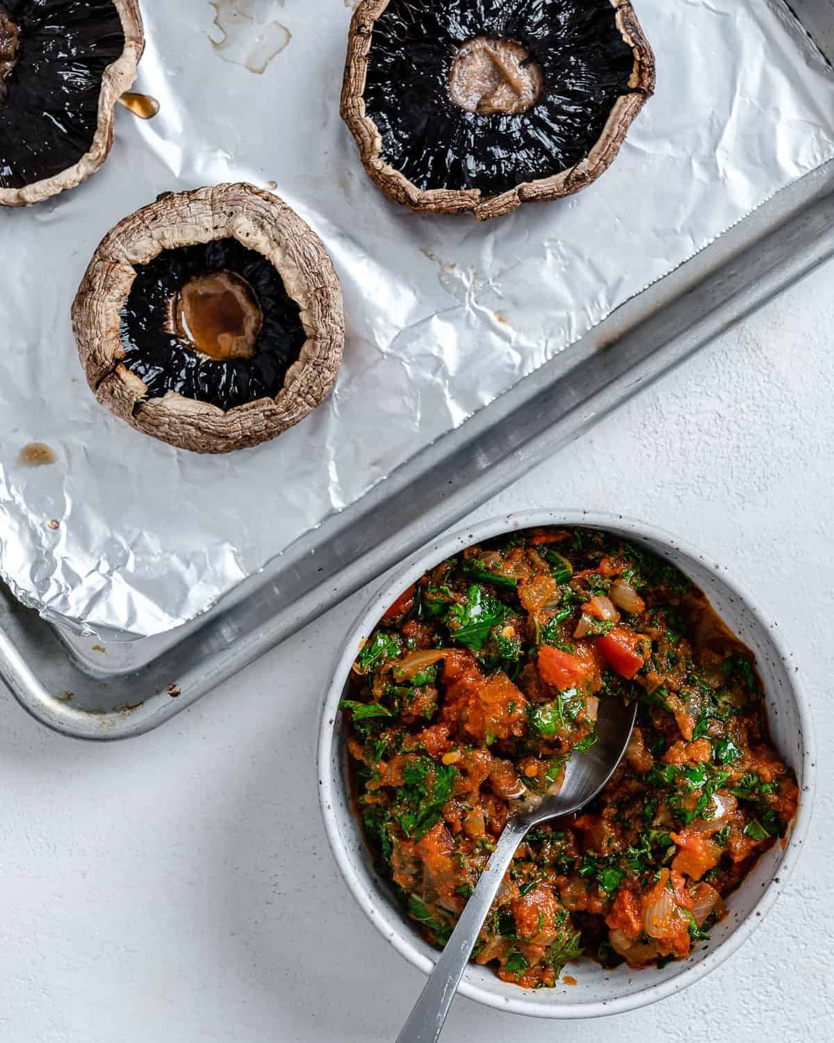 pan of sauce alongside baking tray of portobello mushrooms against a white background