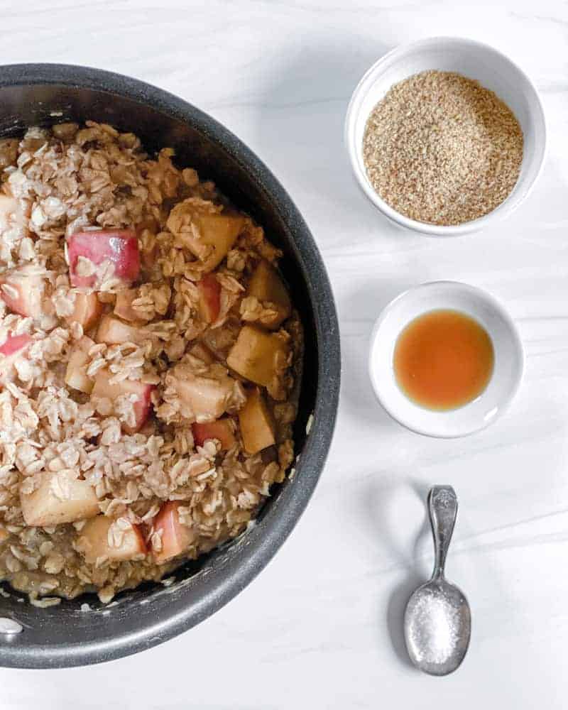 Cooked apple cinnamon oatmeal in a saucepan with the other ingredients behind it.