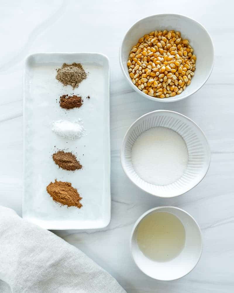 ingredients for chai popcorn measured out on a white tray and white bowls against marble background