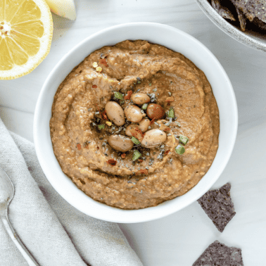 completed Spiced Refried Bean Dip in a bowl against a white background with ingredients spread in the back