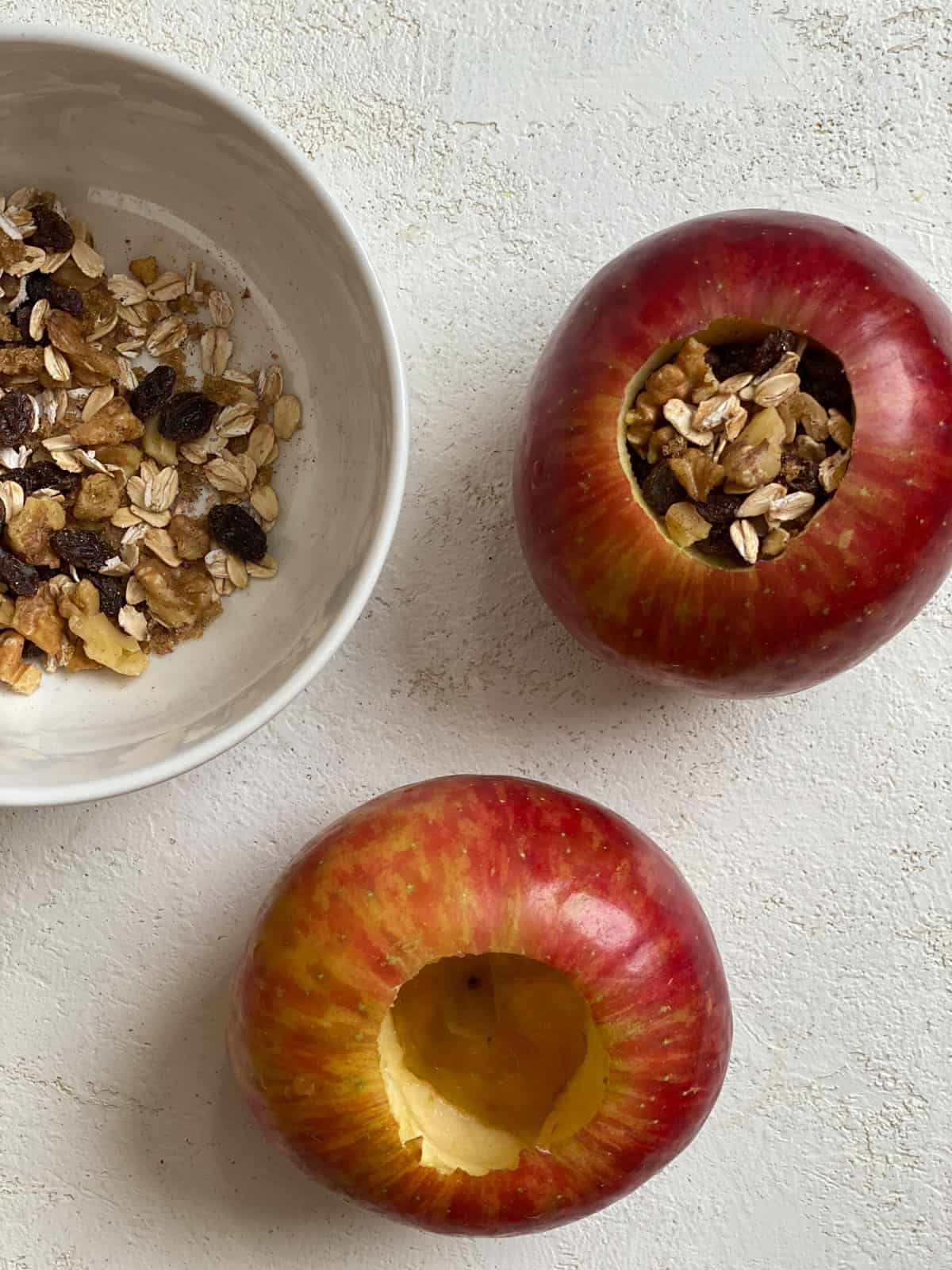 one apple with filling inside alongside a filling-less apple alongside a bowl of filling for stuffed baked apples against a white surface
