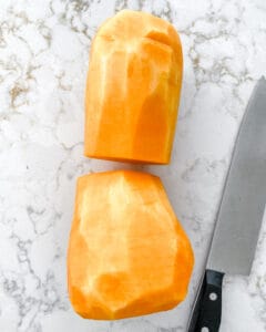 peeled and halved butternut squash with knife against a white marble background