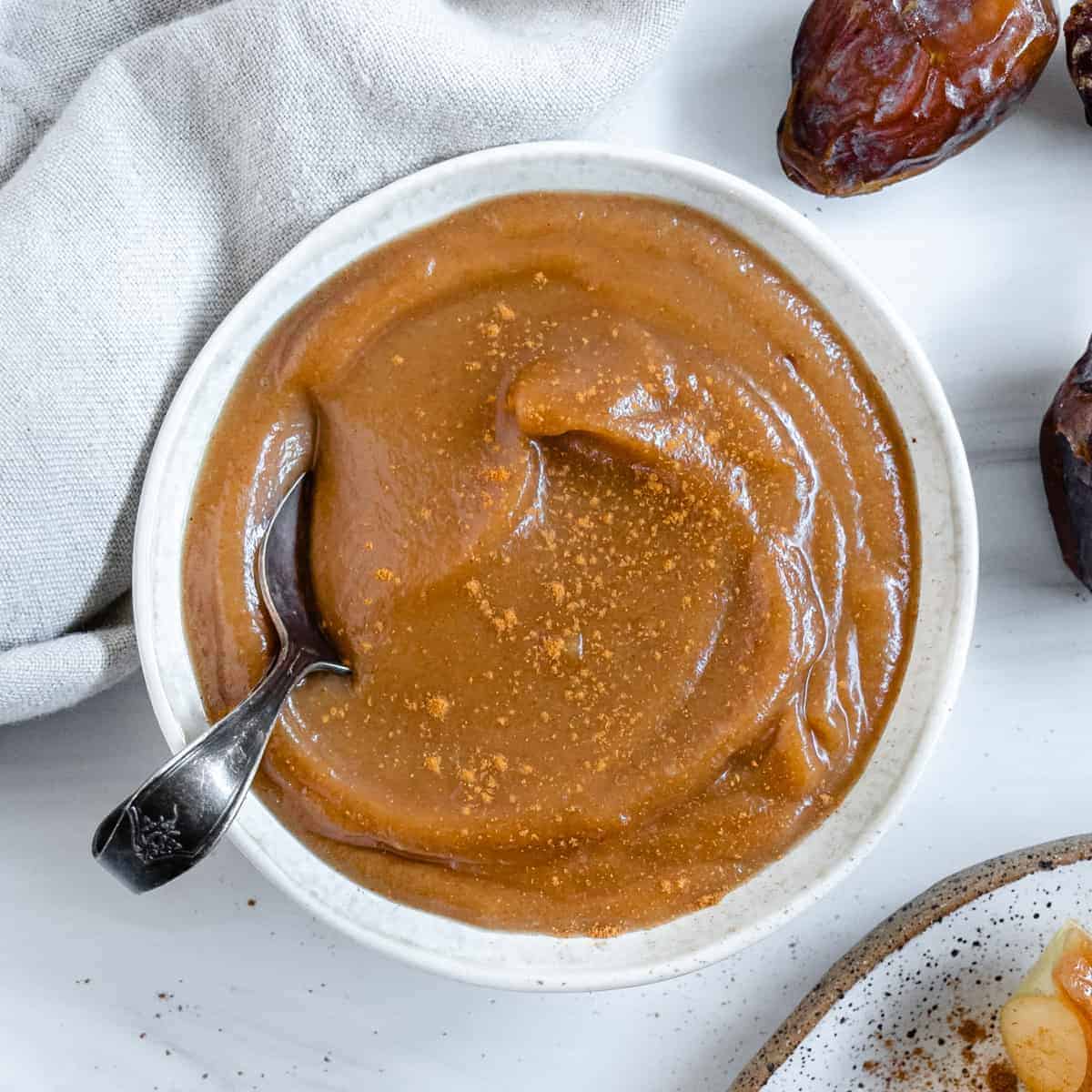 finished homemade caramel sauce in a white bowl with a spoon and dates in the background