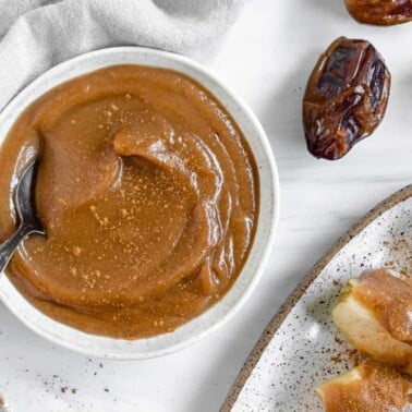 finished homemade caramel sauce in a white bowl with a spoon and dates in the background
