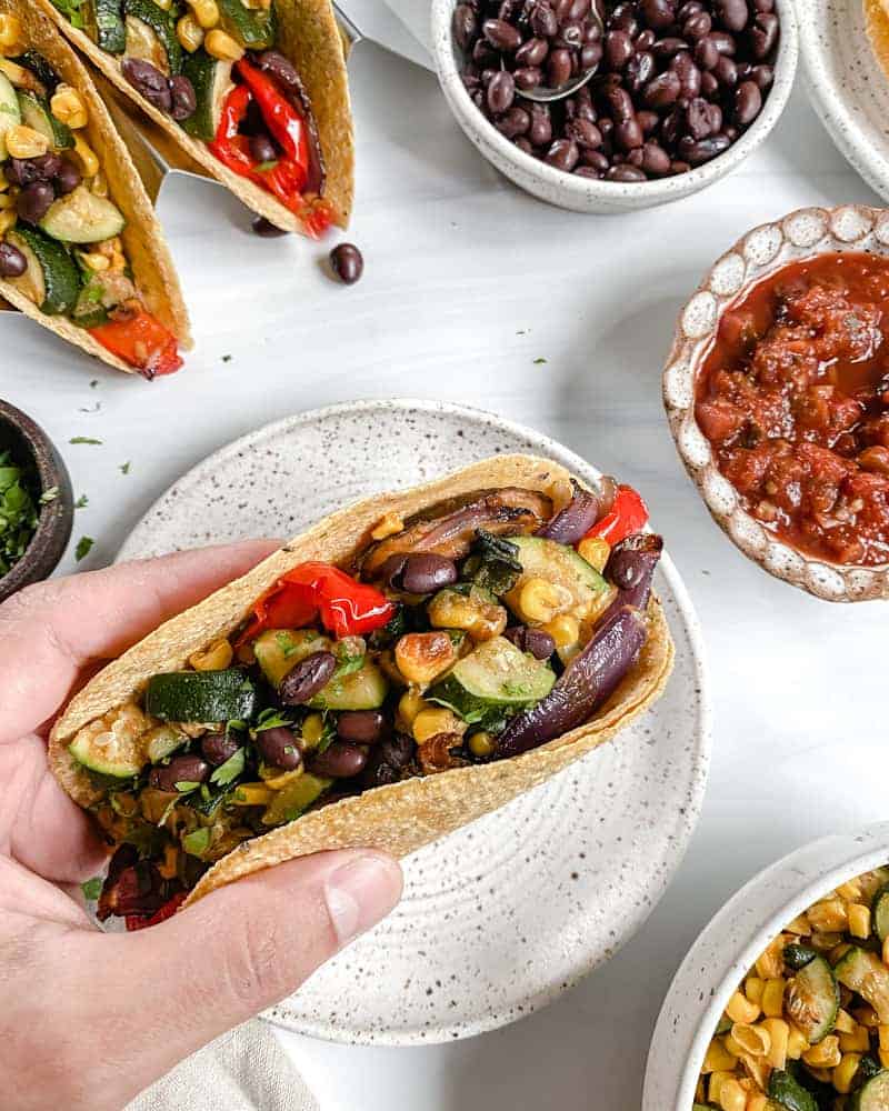 Vegan Roasted Veggie Tacos in a white plate with ingredients in the background against a white surface