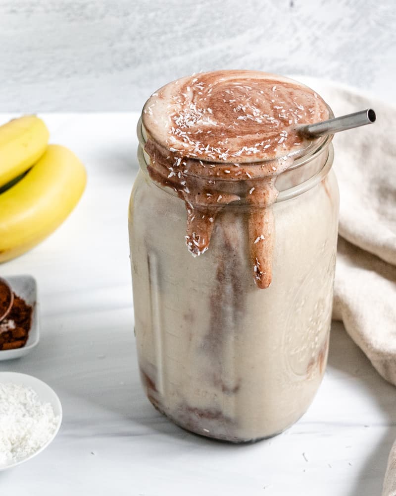 completed C،colate Peanut Butter Banana Smoothie in a jar with ingredients in the background