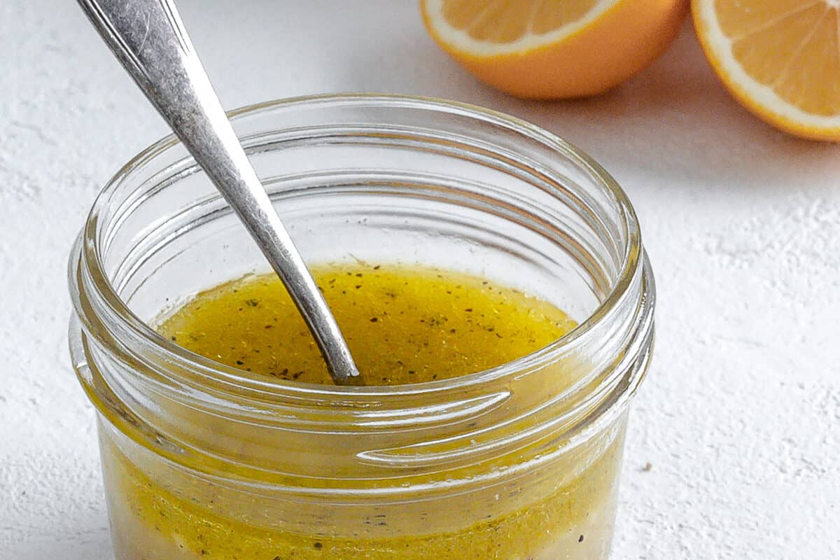 completed lemon dressing in a glass jar with a spoon in it and ingredients in the white background