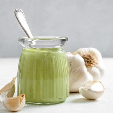 completed Creamy Vegan Garlic Sauce in a glass jar with garlic in the background