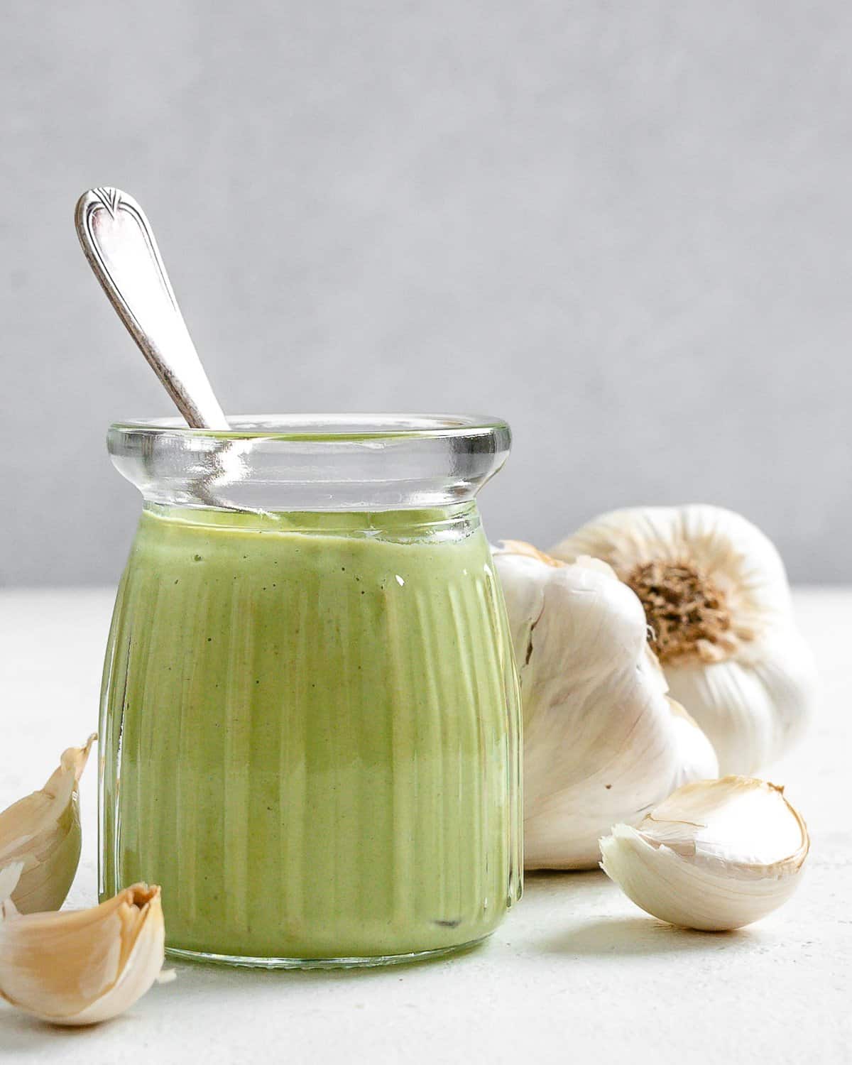 completed Creamy Vegan Garlic Sauce in a glass jar with garlic in the background