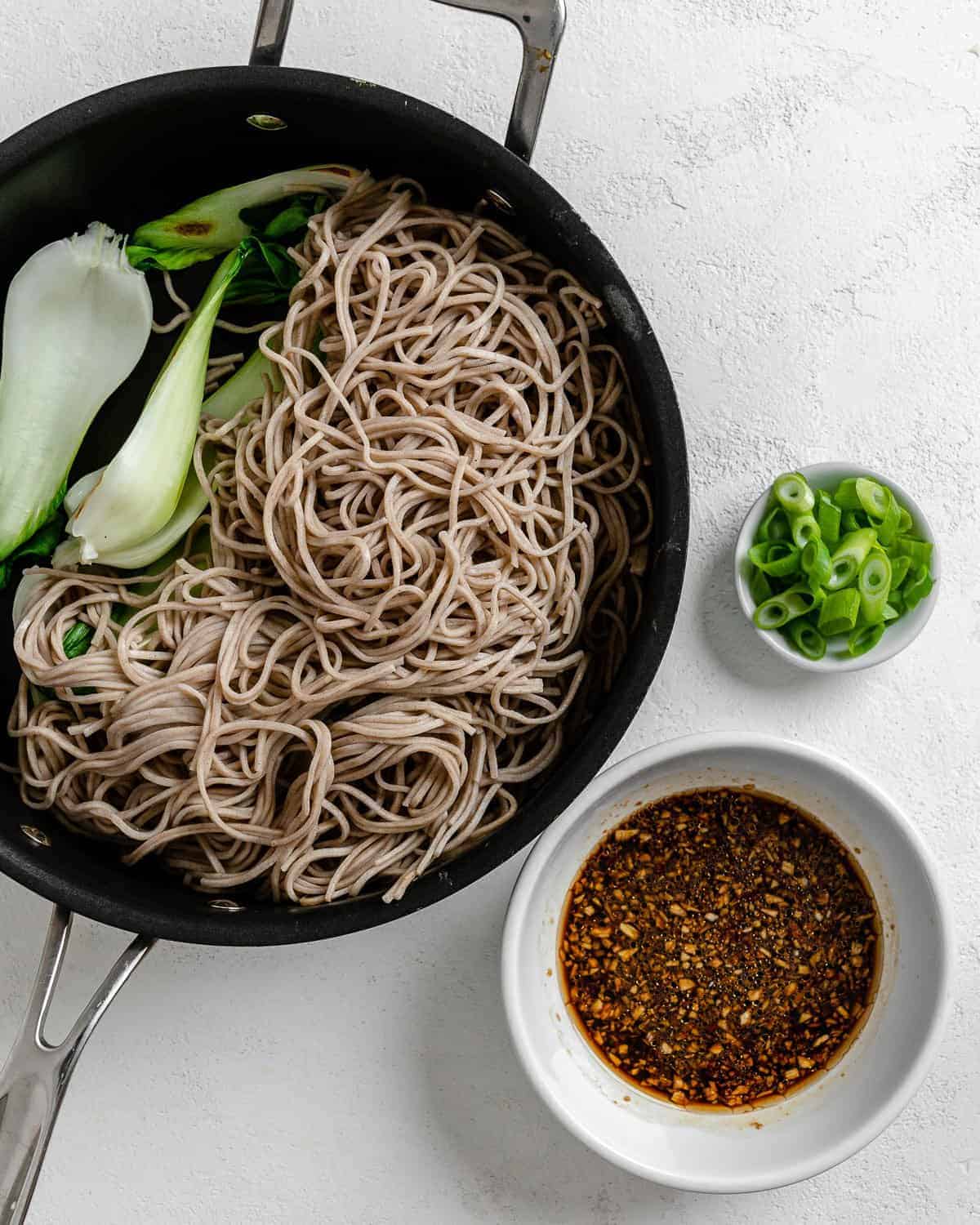 process of addition of soba noodles with veggies in black pan against a white background
