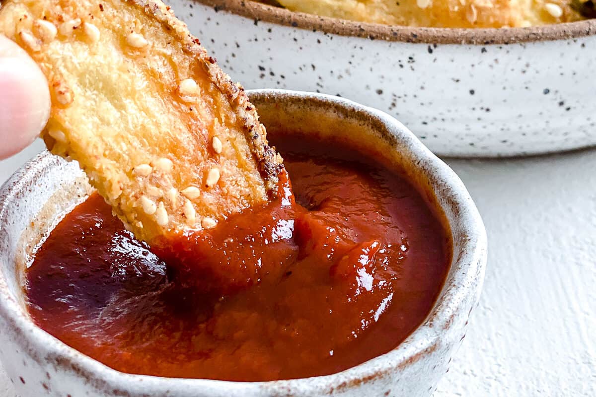 one oven baked sesame fry being dipped into a small bowl of ketchup with fries in the background