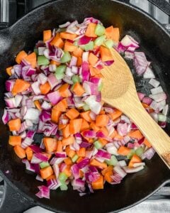 process of sauteing the mirepoix in a black skillet with wooden spoon