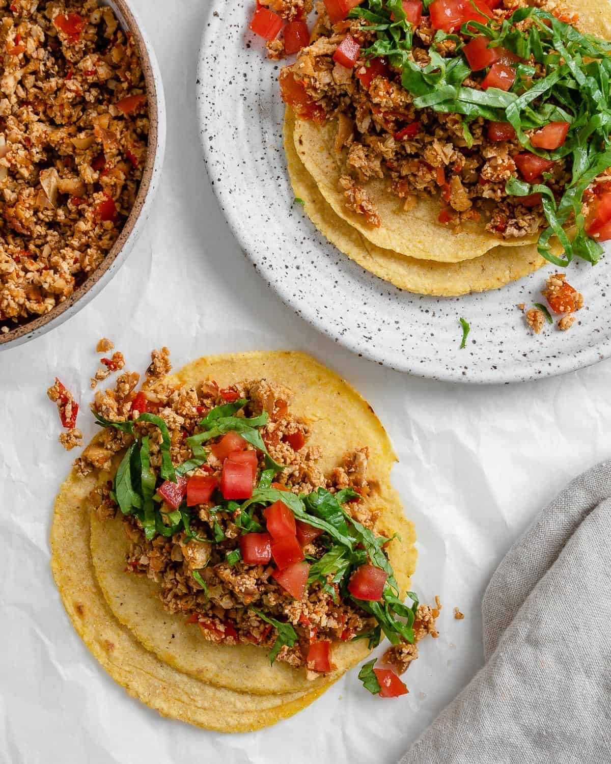 completed tofu tacos on two corn tortillas against a white surface with a plate of tacos in the background