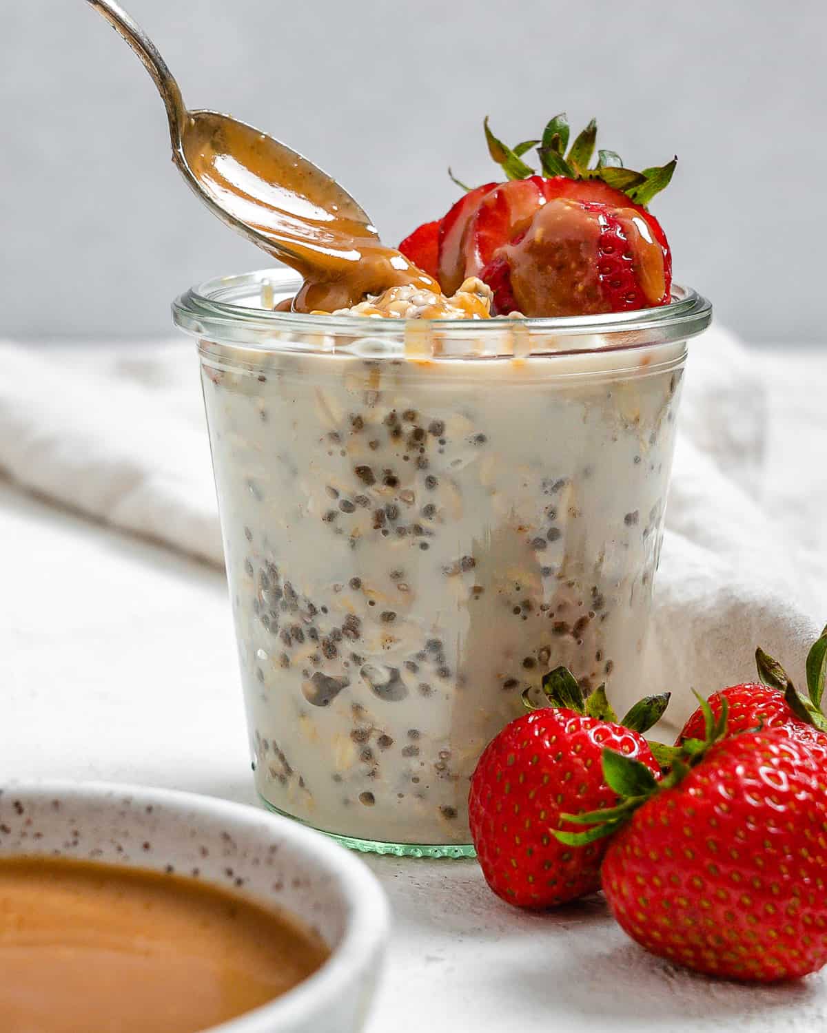completed Salted Caramel Overnight Oats in a clear cup with ingredients against a white background