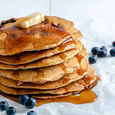 completed stack of Blueberry Pancakes with maple syrup being drizzled on the stack with blueberries scattered against a white background