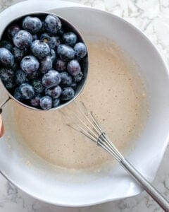 process of adding blueberries to batter in a white bowl