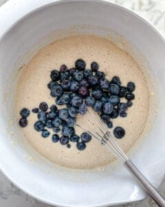process of adding blueberries to batter in a white bowl