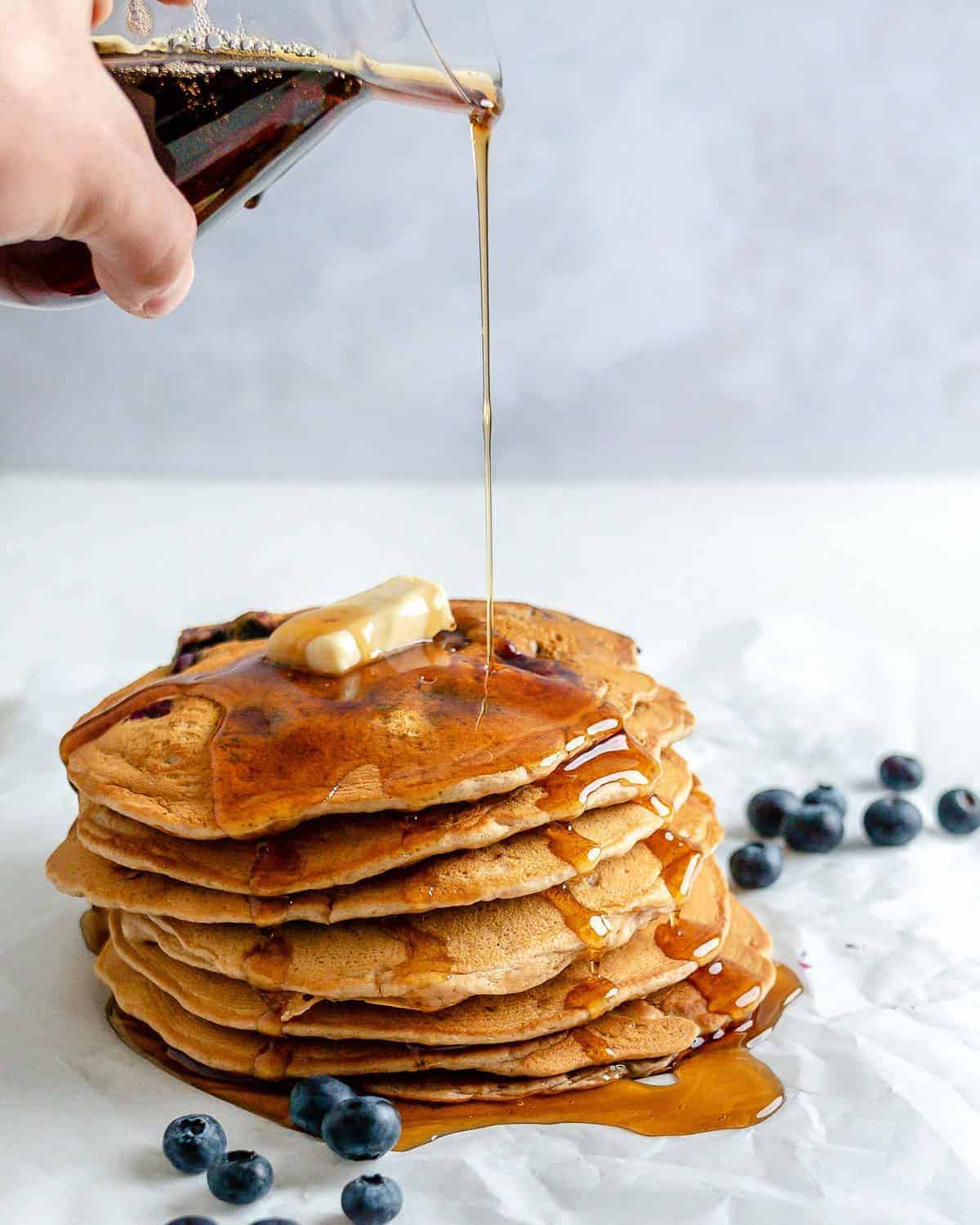 completed stack of Blueberry Pancakes with maple syrup being drizzled on the stack with blueberries scattered against a white background