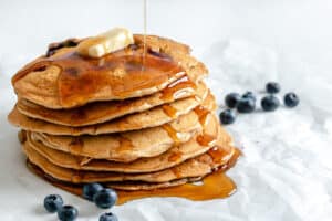 completed stack of Blueberry Pancakes with maple syrup being drizzled on the stack with blueberries scattered against a white background