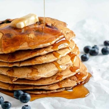 completed stack of Blueberry Pancakes with maple syrup being drizzled on the stack with blueberries scattered against a white background