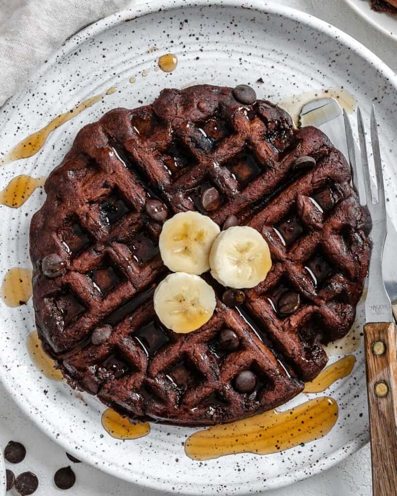 completed waffle on a white plate against a white background with syrup scattered on waffle