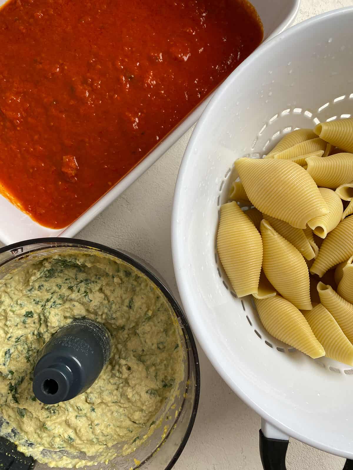 marinara sauce in baking tray alongside with colander of shells and food processor with stuffed shells mixture inside